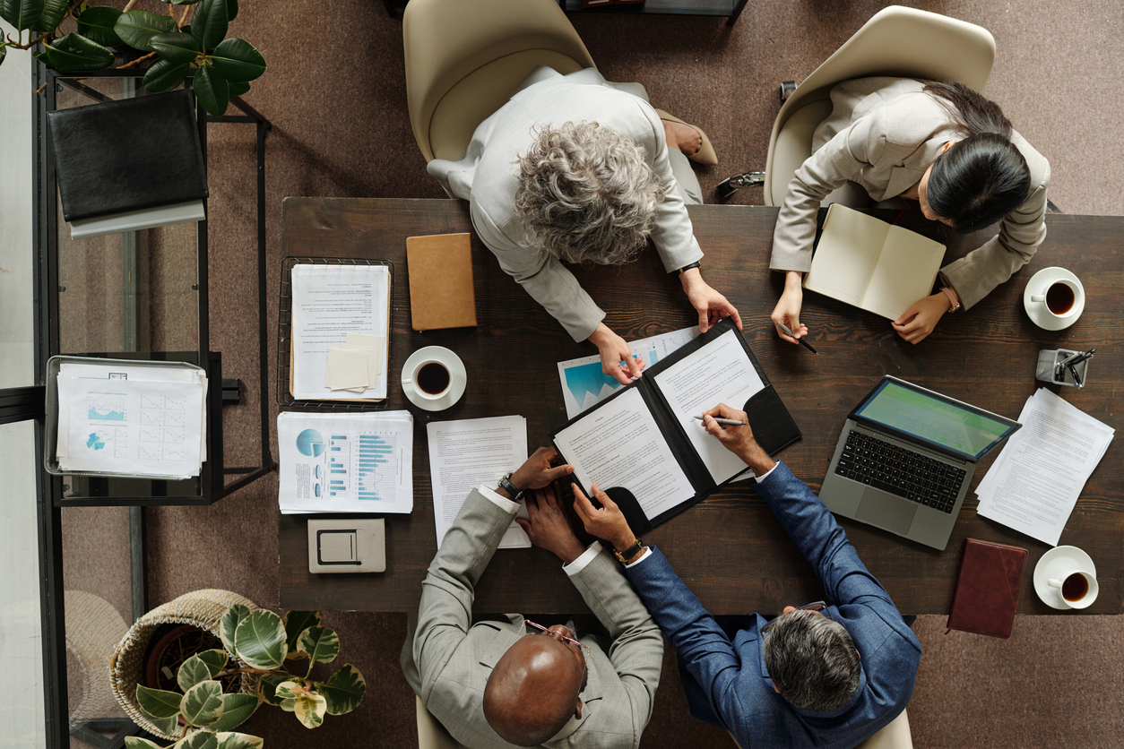 Top-down view of four business professionals collaborating over documents, charts, and a laptop at a wooden conference table.